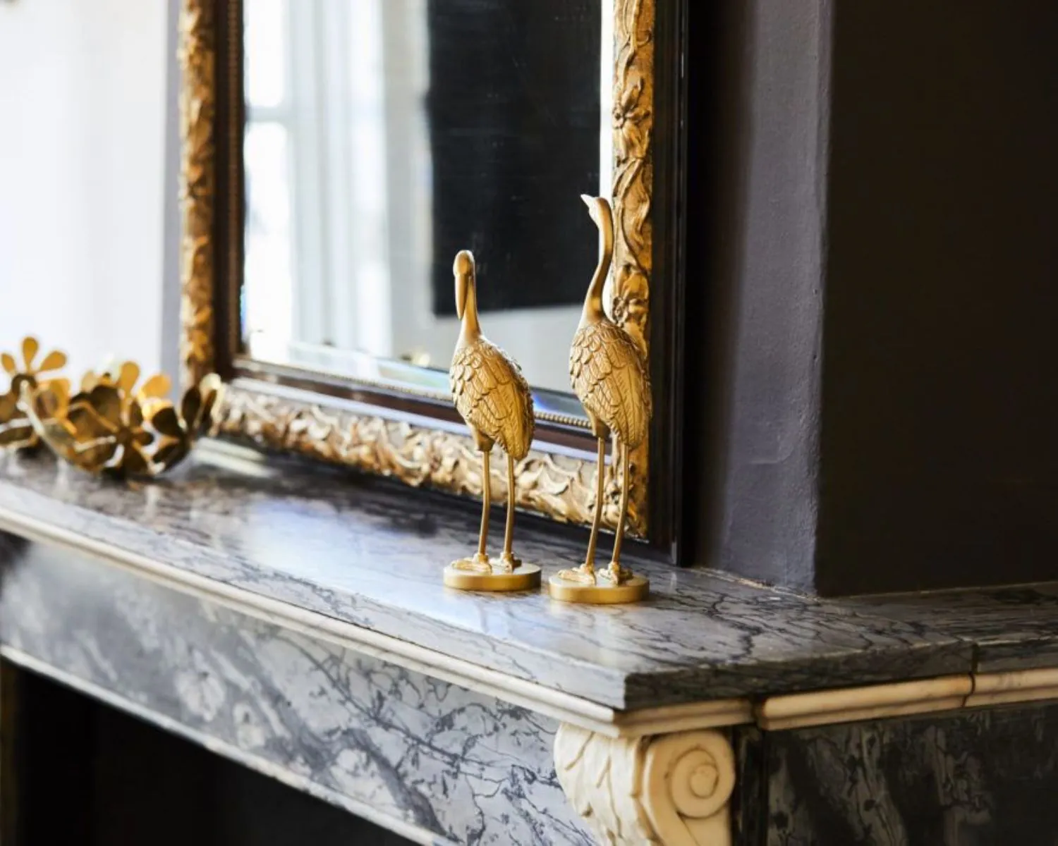 Twee gouden sierlijke vogelbeelden op een marmeren schouw met een grote sierlijstspiegel op de achtergrond.