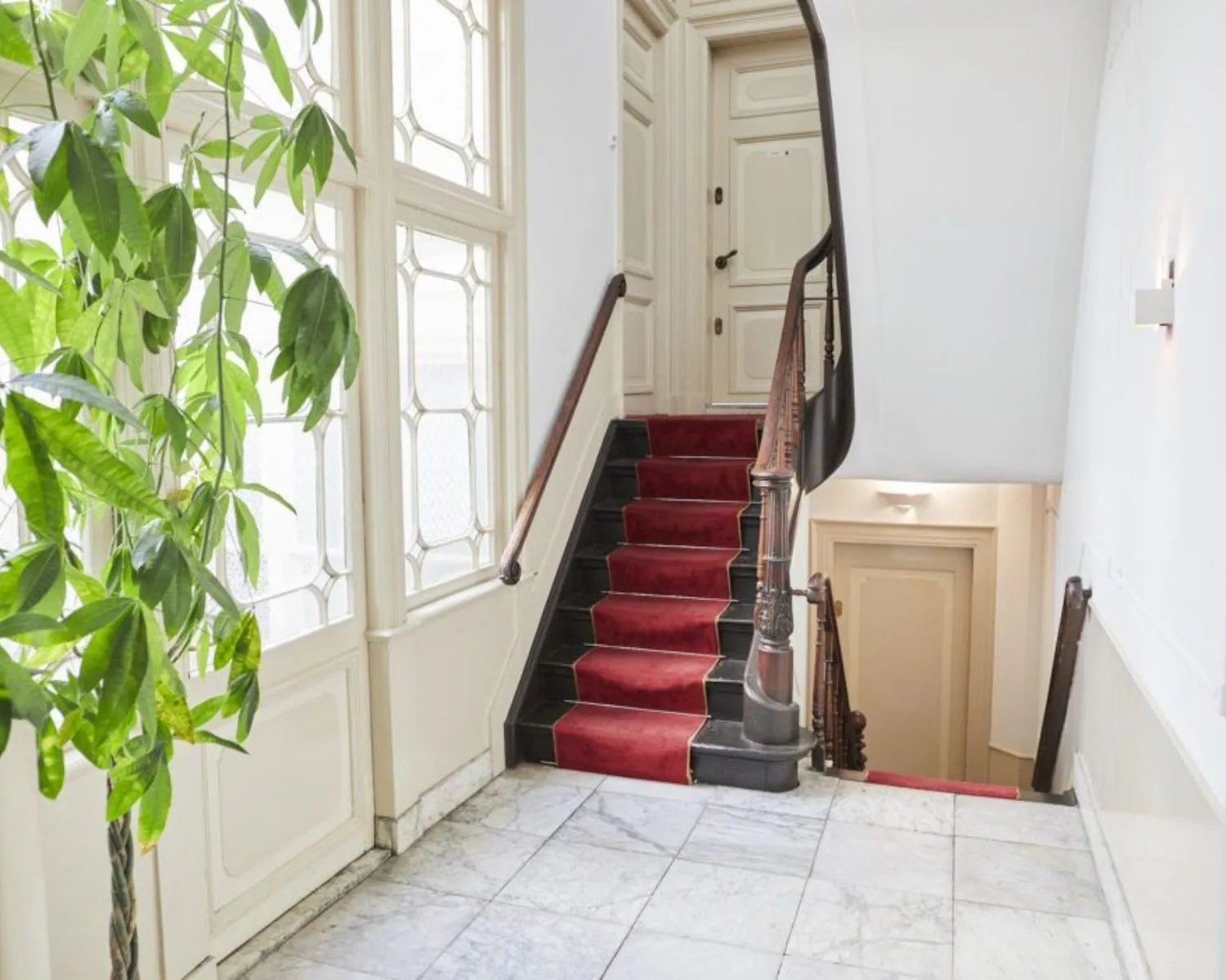 Trap met rode loper in een klassiek trappenhuis met marmeren vloer en grote raampartij.