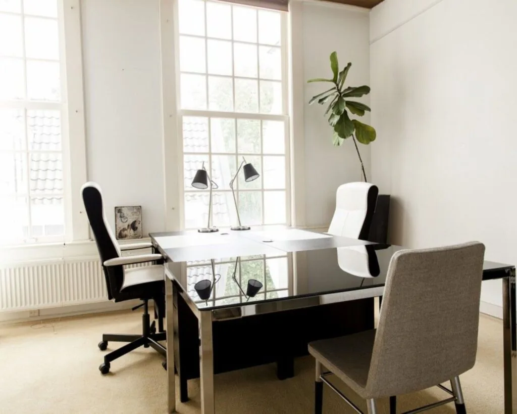 Modern en licht kantoor met grote ramen, een glazen bureau, drie stoelen en een kamerplant.