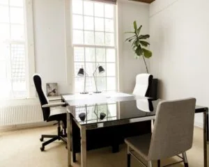 Modern en licht kantoor met grote ramen, een glazen bureau, drie stoelen en een kamerplant.