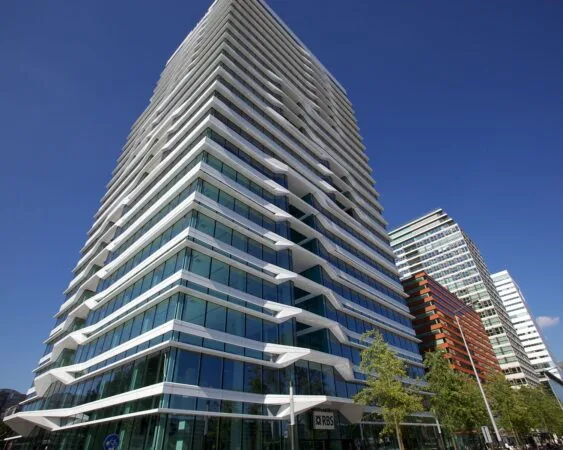 Modern kantoorgebouw met glazen gevel en witte, golvende balkonlijnen in een stedelijke omgeving onder een blauwe lucht.