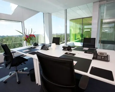 Modern, licht kantoor met glazen wanden, uitgerust met bureaustoelen, telefoons en een vaas met bloemen op een witte vergadertafel.