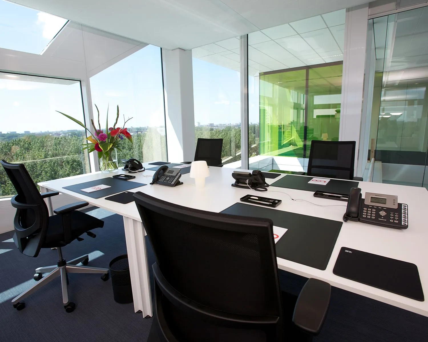 Modern, licht kantoor met glazen wanden, uitgerust met bureaustoelen, telefoons en een vaas met bloemen op een witte vergadertafel.