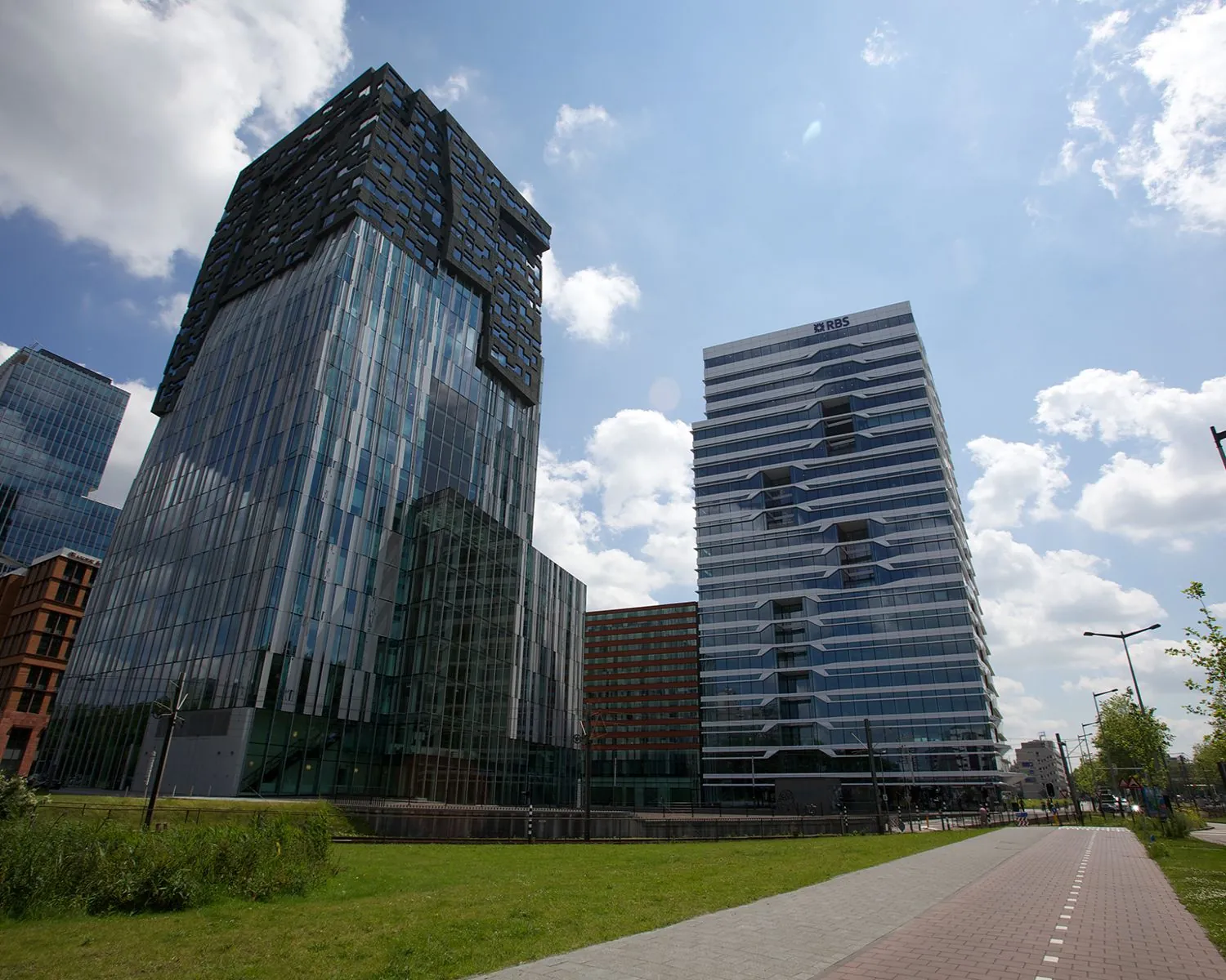 Twee moderne kantoorgebouwen, waaronder het RBS-gebouw, aan een fietspad met gras en blauwe lucht op de achtergrond.
