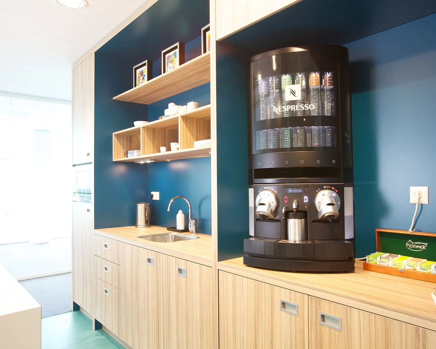 Moderne kantoorkeuken met een Nespresso koffiemachine, houten kasten en open planken met servies.