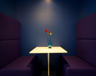 Wit tafeltje met een blauwe vaas en rode bloem tussen twee paarse zitbanken in een blauwe nis.