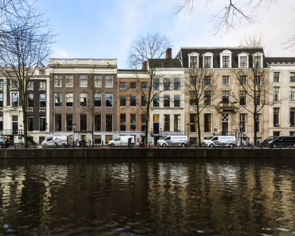 Rij statige grachtenpanden aan een Amsterdamse gracht met geparkeerde bestelbusjes op de kade.