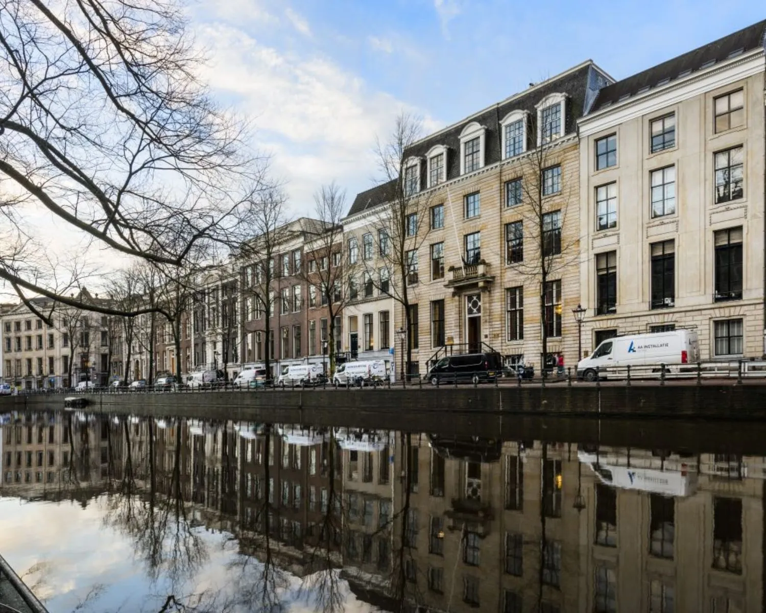 Rijtje statige grachtenpanden weerspiegelt in het water van een Amsterdamse gracht op een heldere dag.