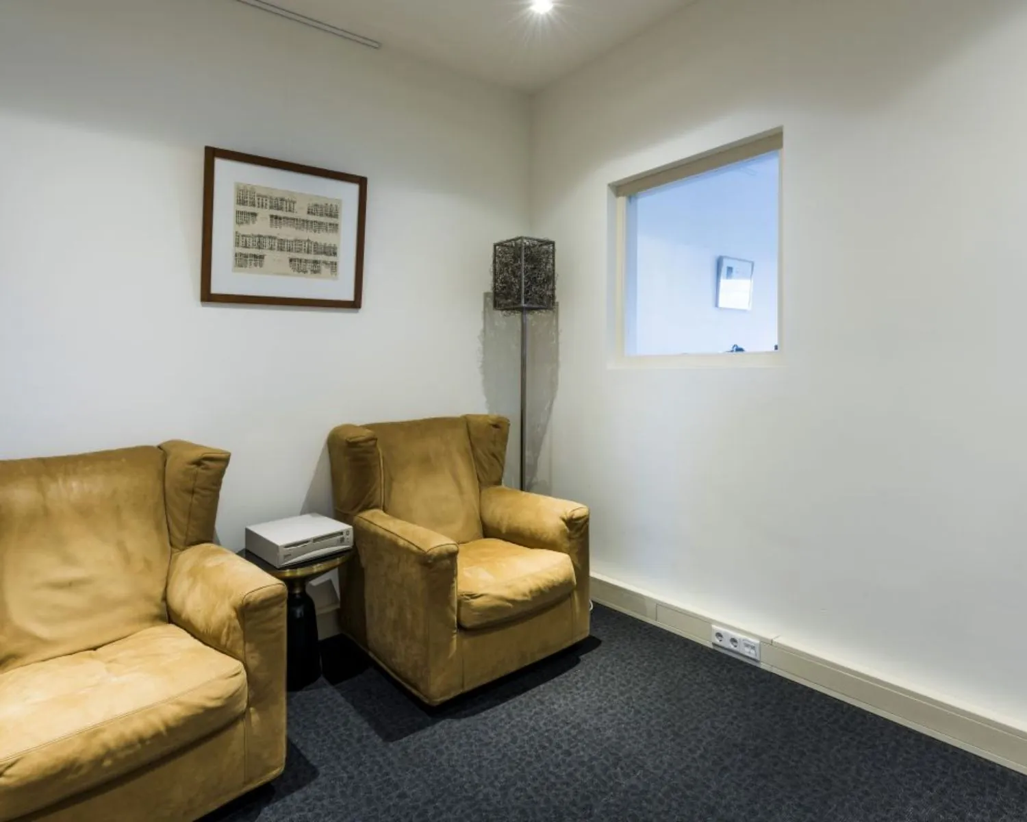 Twee gele leunstoelen staan in een minimalistische wachtruimte met witte muren, een schilderij en een klein raam.