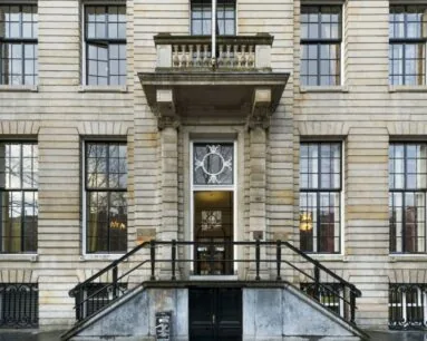 Hoofdingang van een statig, klassiek gebouw met symmetrische ramen en een trap met smeedijzeren leuning.