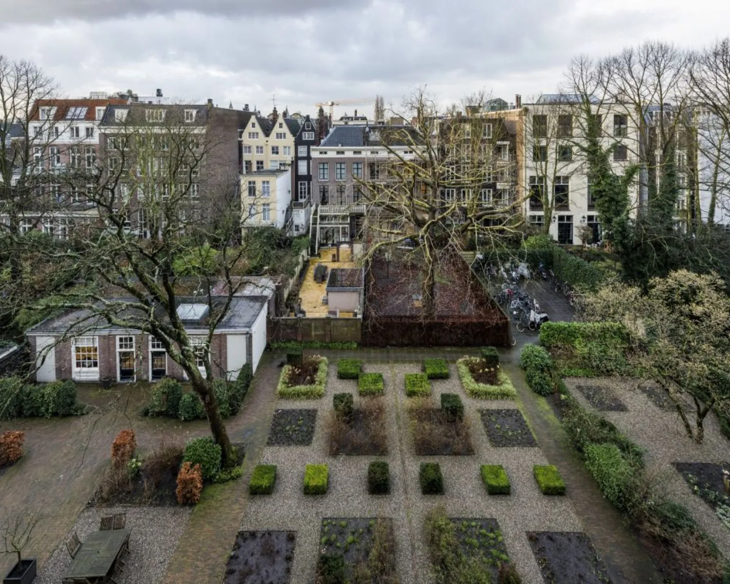 Luchtfoto van een verzorgde stadstuin met grindpaden, struiken en bomen, omgeven door hoge herenhuizen.