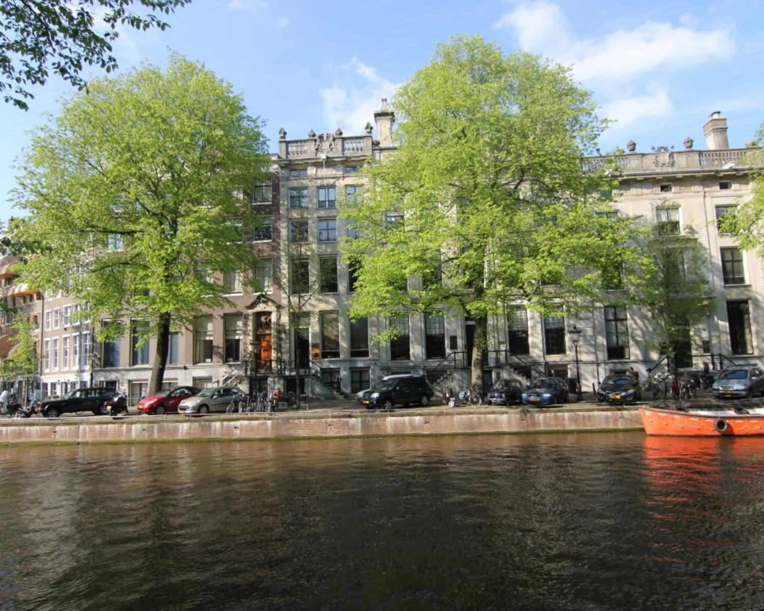 Grachtepandengevels met groene bomen aan een Amsterdamse gracht op een zonnige lentedag.