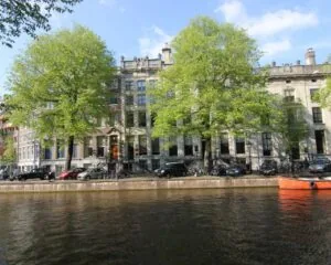 Grachtepand aan het water in Amsterdam met geparkeerde auto’s en een oranje boot op de voorgrond.