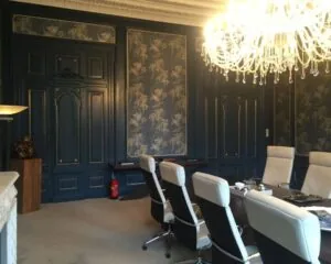 Vergaderruimte met klassieke donkerblauwe lambrisering, een grote kristallen kroonluchter en moderne witte stoelen rond een tafel.