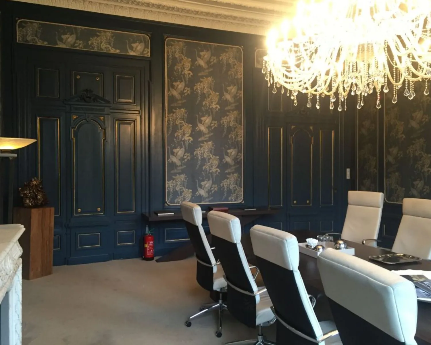 Vergaderruimte met klassieke donkerblauwe lambrisering, een grote kristallen kroonluchter en moderne witte stoelen rond een tafel.