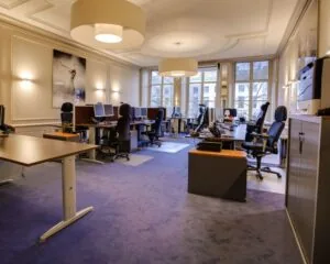 Ruim en modern kantoor met meerdere werkplekken, bureaustoelen en computers in een lichte omgeving met grote ramen.