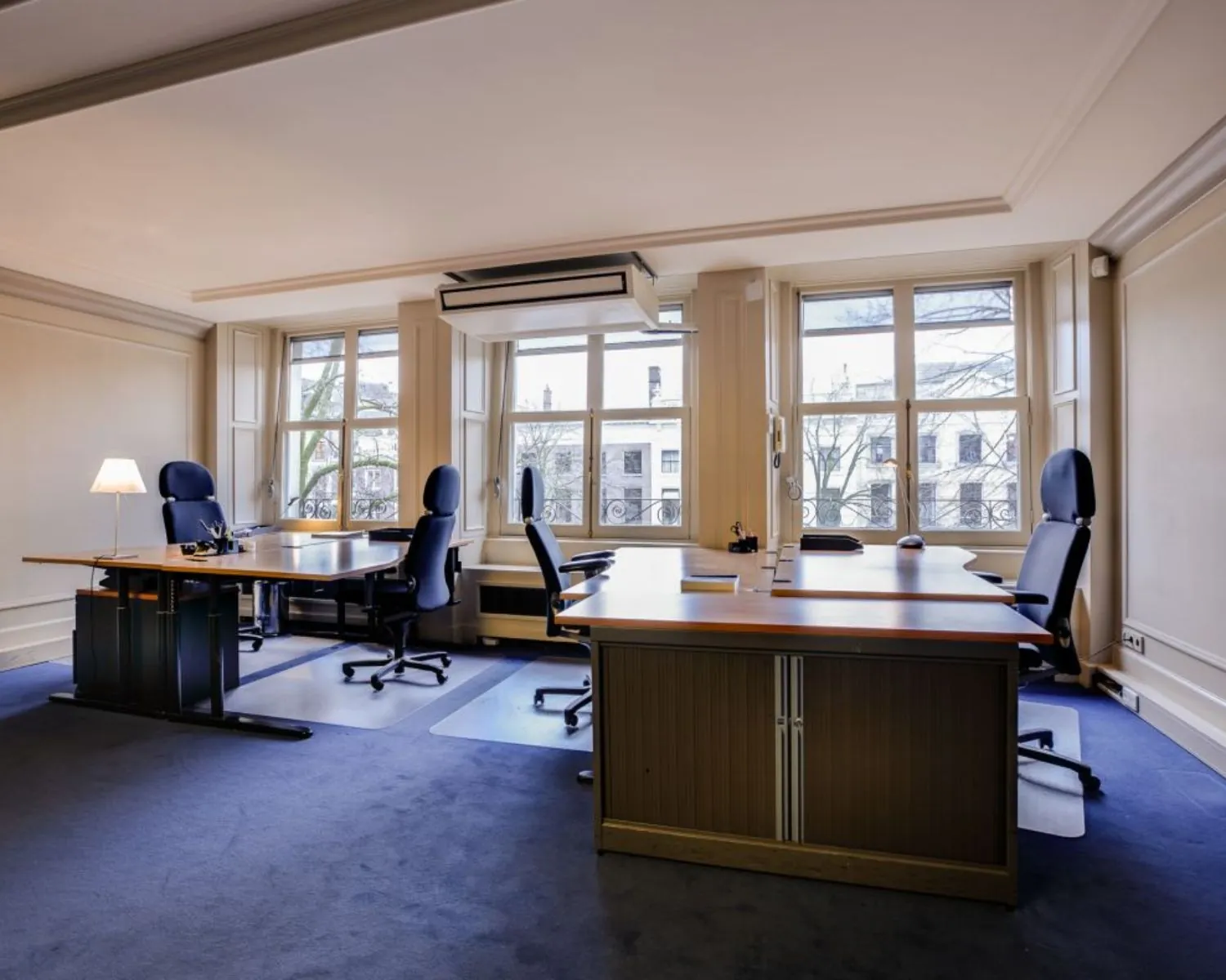 Ruim en licht kantoor met vier bureaus en ergonomische bureaustoelen bij grote ramen.