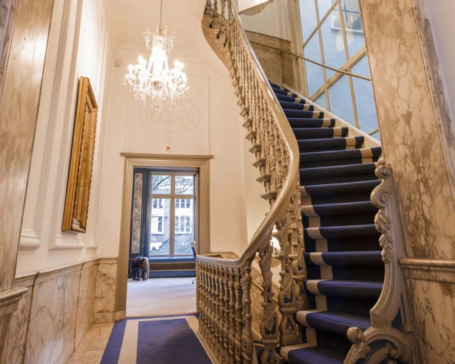Monumentaal trappenhuis met blauwe loper, marmeren muren en een kristallen kroonluchter.