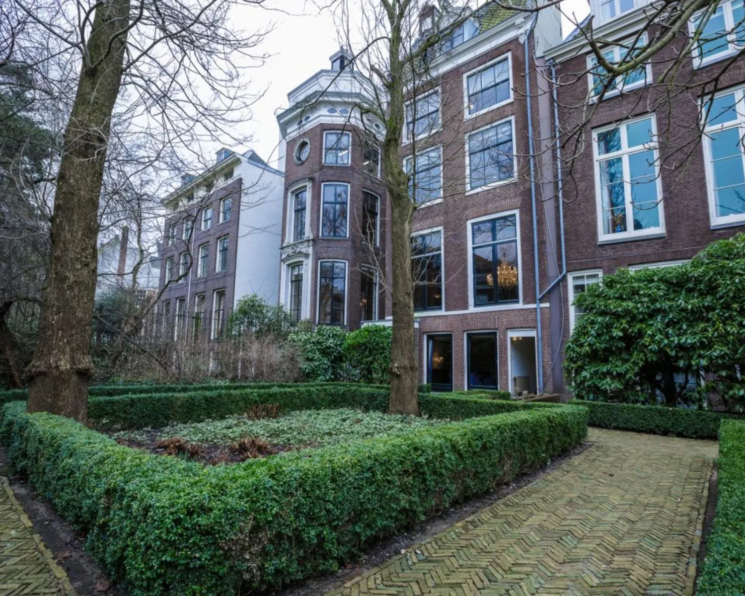 Achterzijde van een klassiek herenhuis met hoge ramen, omringd door een formele tuin met buxushaag en klinkerpad.