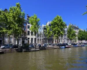 Rij traditionele grachtenpanden met bomen en boten langs een Amsterdamse gracht op een zonnige dag.