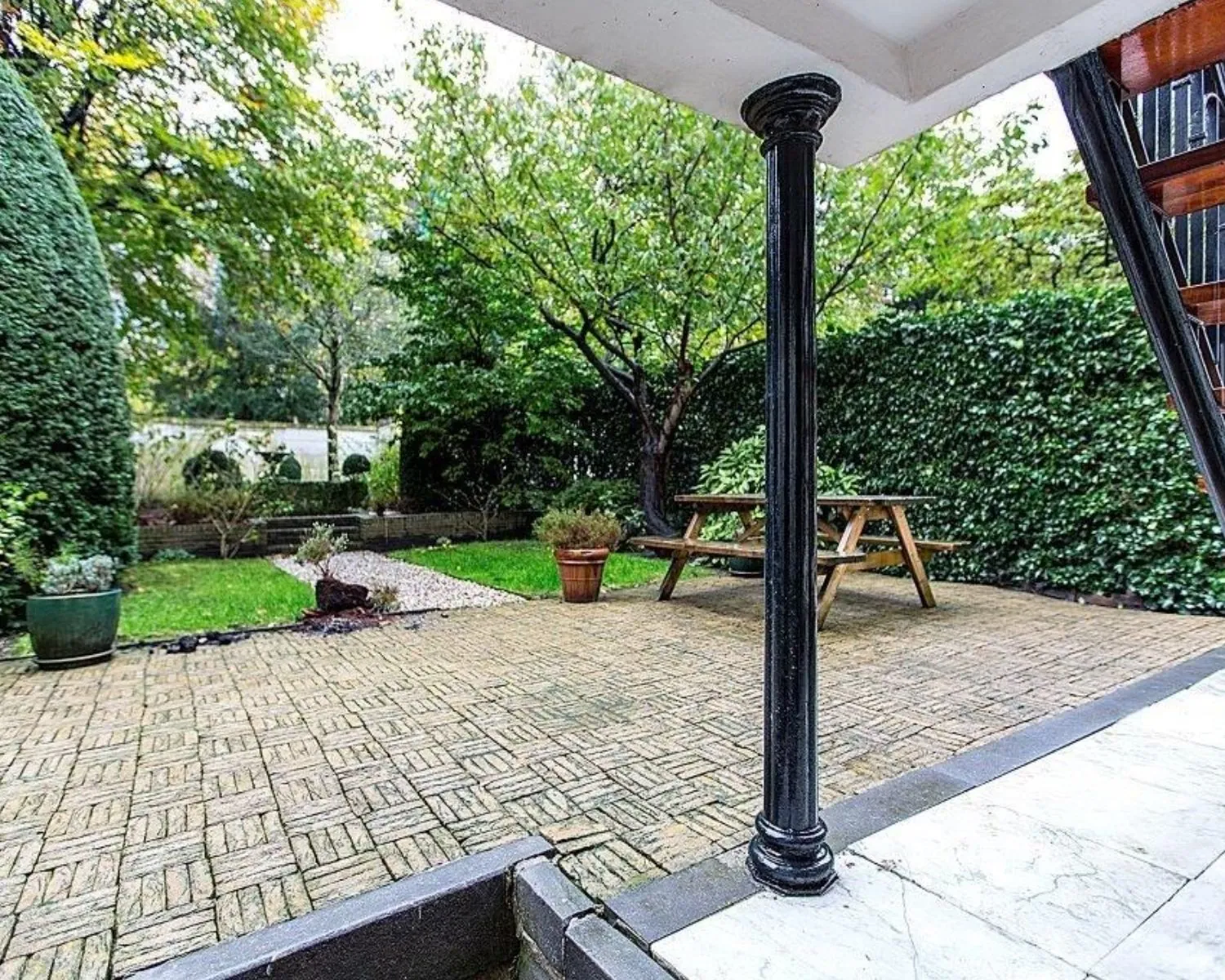 Overdekt terras met zwart metalen zuil, uitzicht op betegelde tuin met picknicktafel en groene begroeiing.