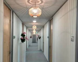 Verlichtte gang met houten deuren, bloemen op sokkels en decoratieve hanglampen aan het plafond.