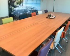 Vergaderruimte met een grote houten tafel, gekleurde stoelen en een strandfoto op de achterwand.