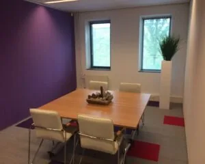 Vergaderruimte met een houten tafel, vier witte stoelen, paarse muur en hoge plantenbak bij het raam.