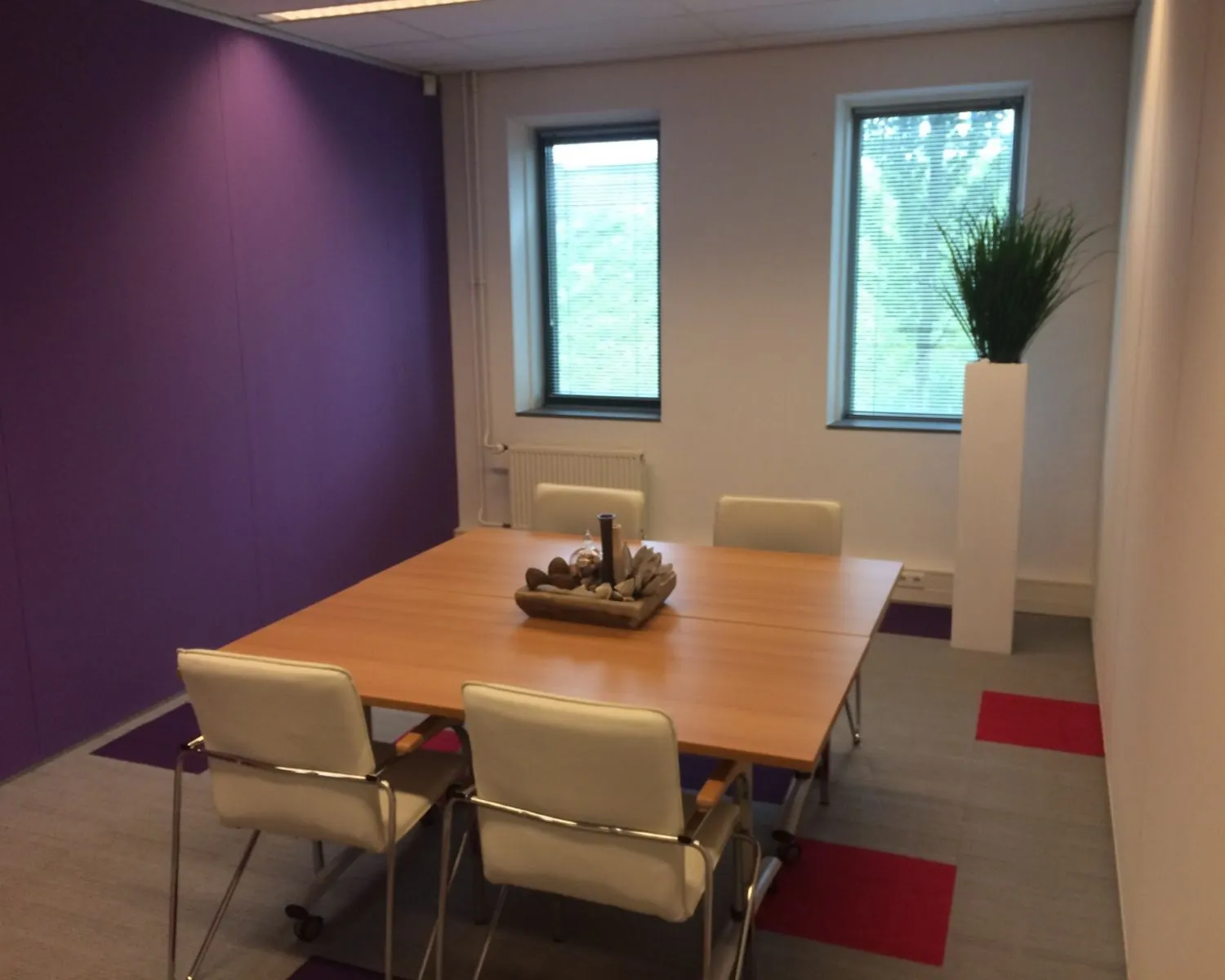 Vergaderruimte met een houten tafel, vier witte stoelen, paarse muur en hoge plantenbak bij het raam.