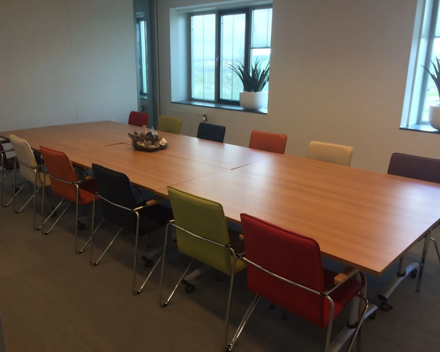 Vergaderruimte met een grote houten tafel en kleurrijke stoelen in een lichte kantooromgeving.