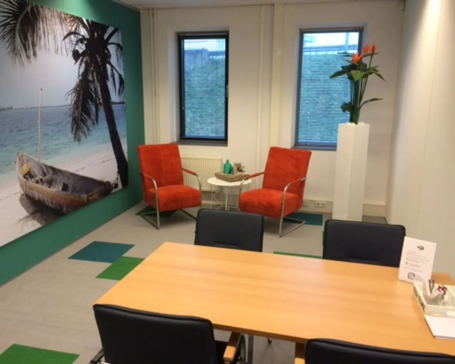 Moderne vergaderruimte met feloranje stoelen, tropisch strandfotobehang en een houten tafel met stoelen.