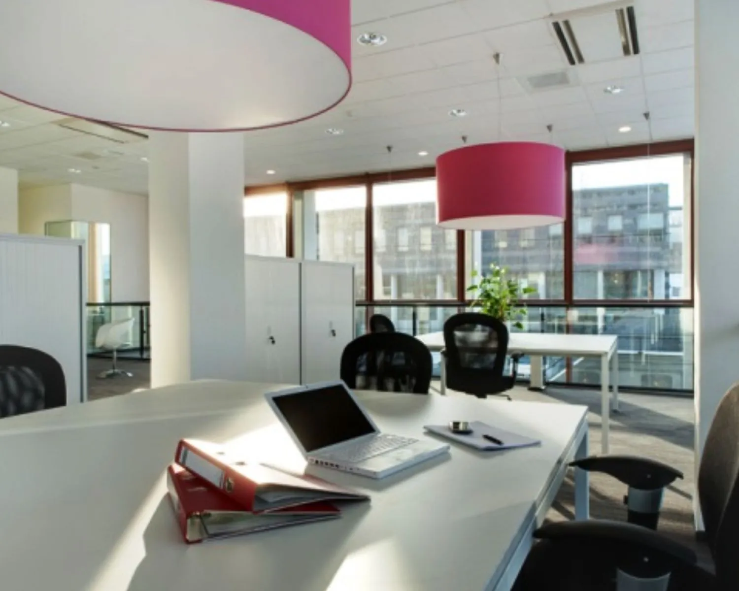 Moderne kantoorruimte met witte bureaus, zwarte stoelen, een open laptop en roze hanglampen.