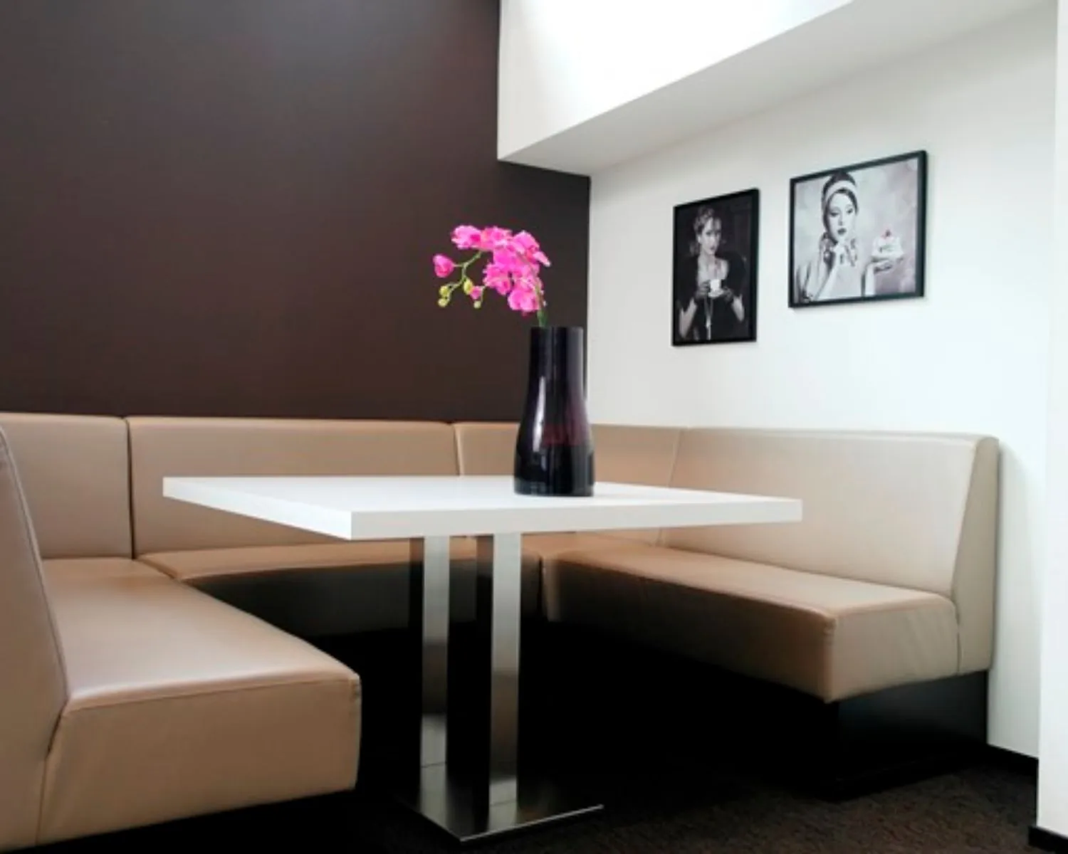 Moderne zithoek met beige banken, een witte tafel, een donkere vaas met roze bloemen en twee zwart-wit portretten aan de muur.