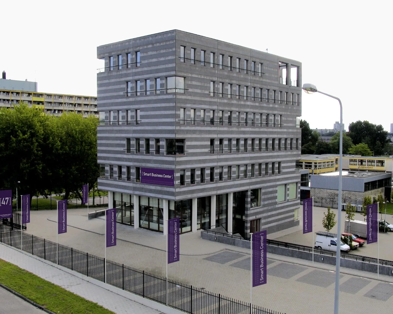 Een modern kantoorgebouw met de naam "Smart Business Center", omgeven door paarse vlaggen en gelegen aan een stadsstraat.