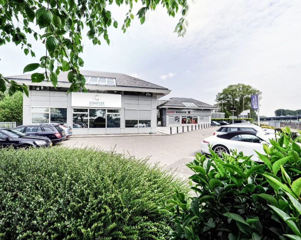 Bedrijventerrein met het pand van Complex Built to Work en Mascot Workwear, omgeven door geparkeerde auto's en groen.
