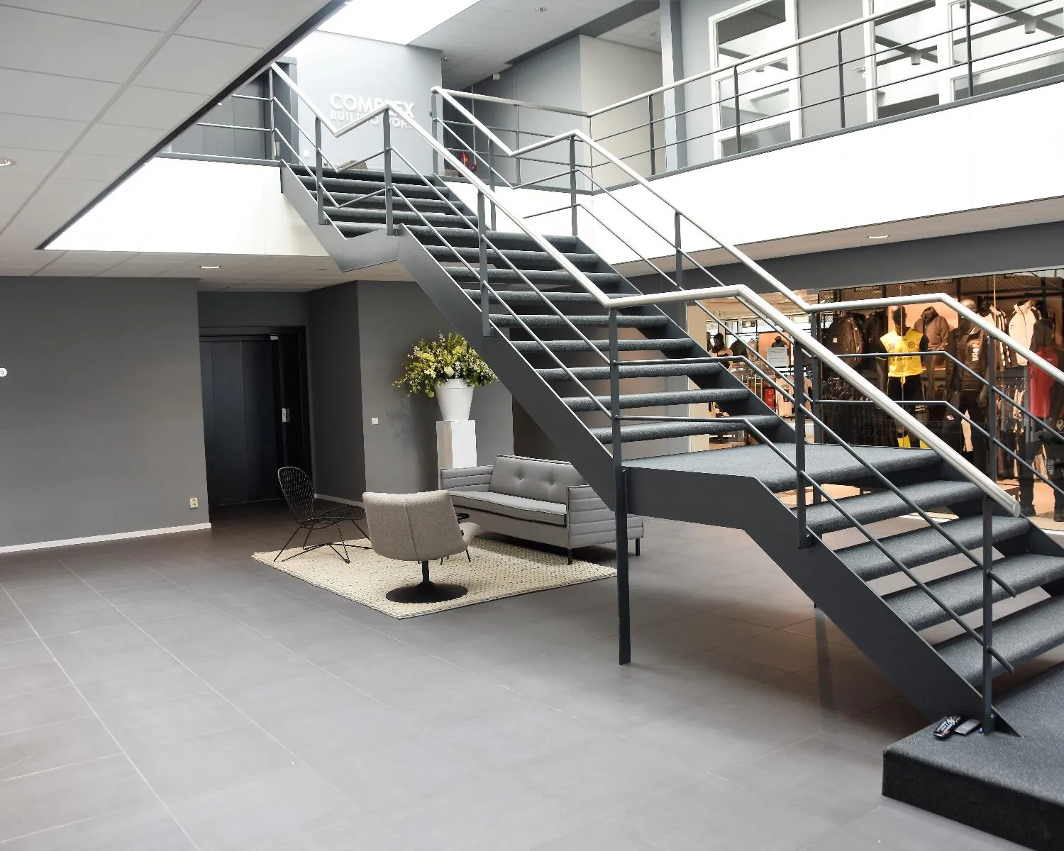 Moderne bedrijfsentree met grijze trap, zitgedeelte en zicht op kledingwinkel op de achtergrond.