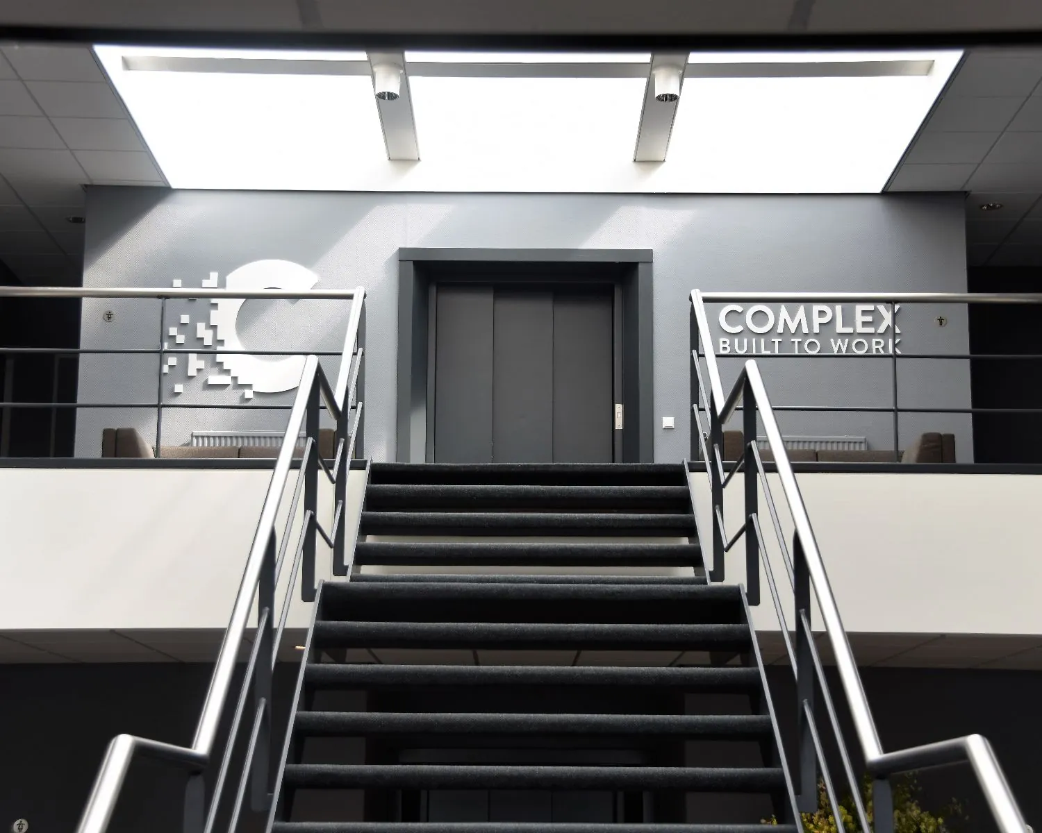 Trap met zwarte treden leidt naar een moderne kantoorverdieping met het logo en de slogan van Complex: "Built to work".