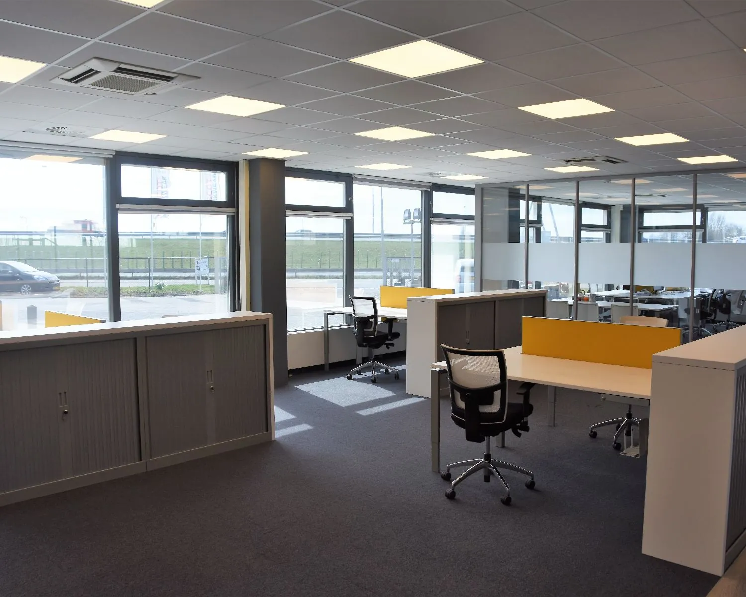 Moderne, open kantoorruimte met bureaus, bureaustoelen en grote ramen die daglicht binnenlaten.