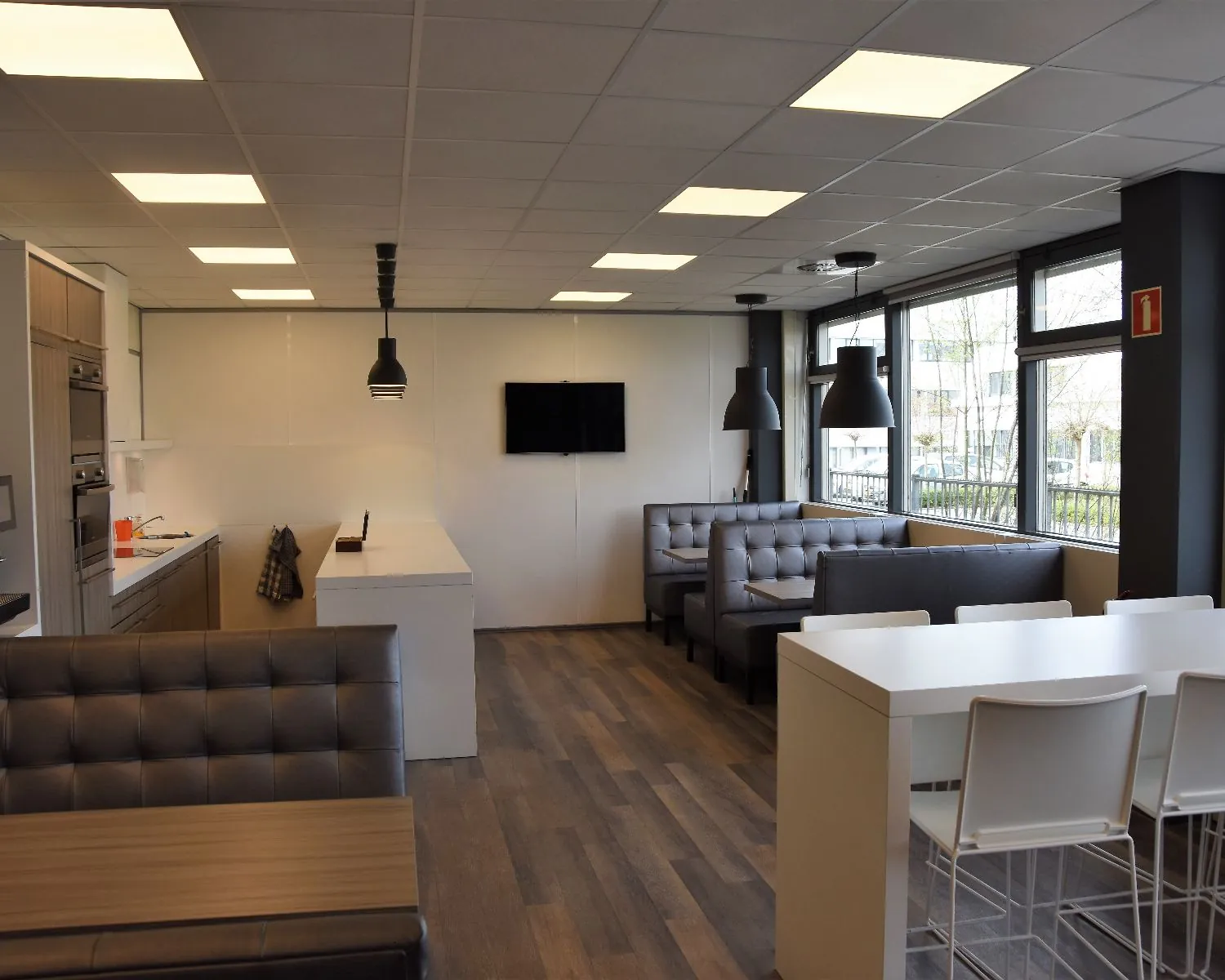 Moderne bedrijfskantine met zithoeken, hoge tafels, een open keuken en grote ramen.