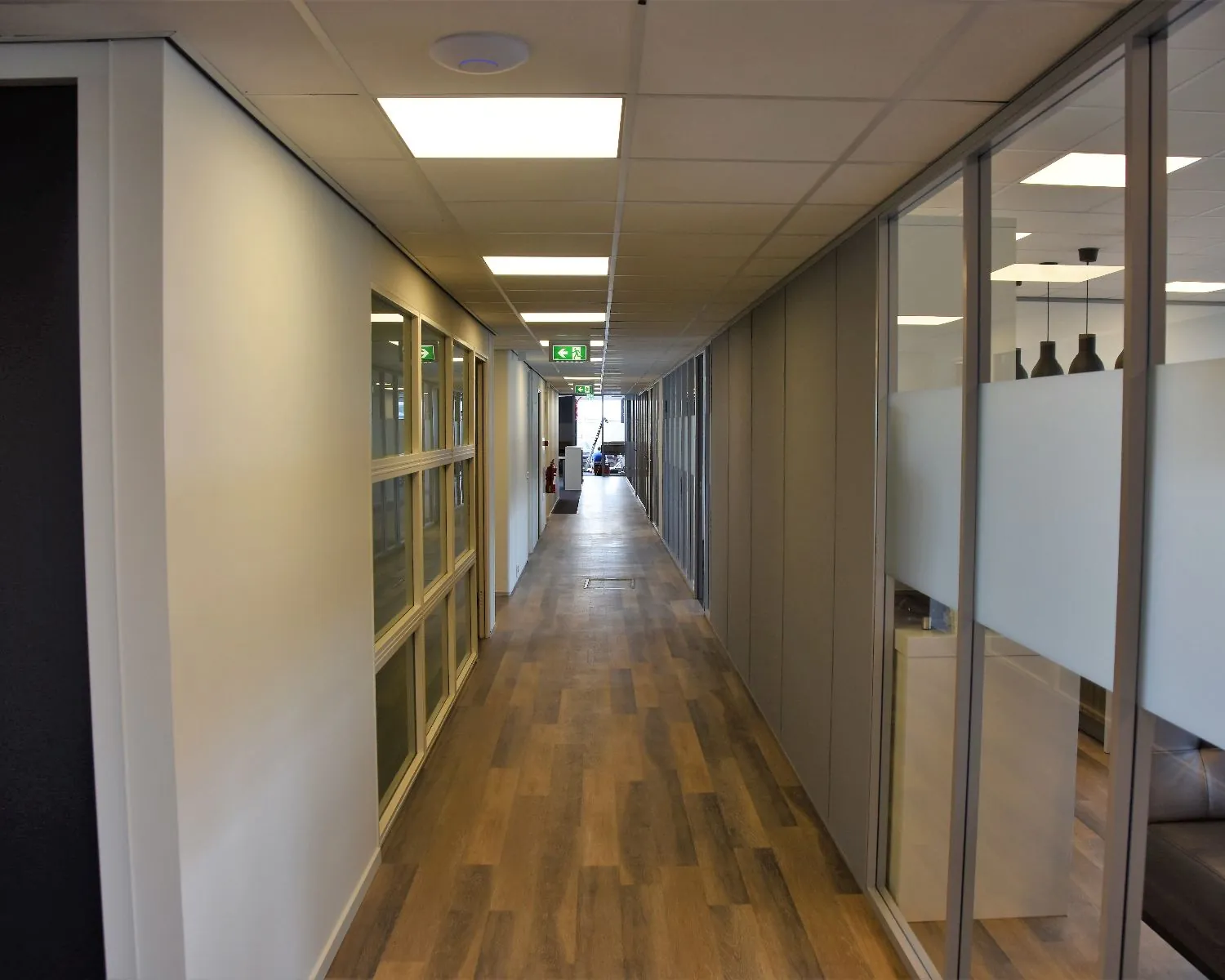 Moderne kantoorhal met houten vloer, systeemplafond en glazen wanden aan beide zijden.