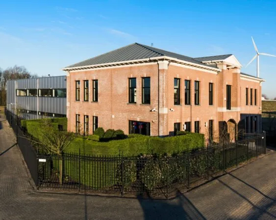 Een modern kantoorgebouw van baksteen en metaal met omheinde tuin en windturbine op de achtergrond.