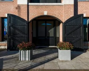 Ingang van een modern gebouw met grote zwarte houten deuren, twee betonnen plantenbakken en een gevel van rode baksteen.