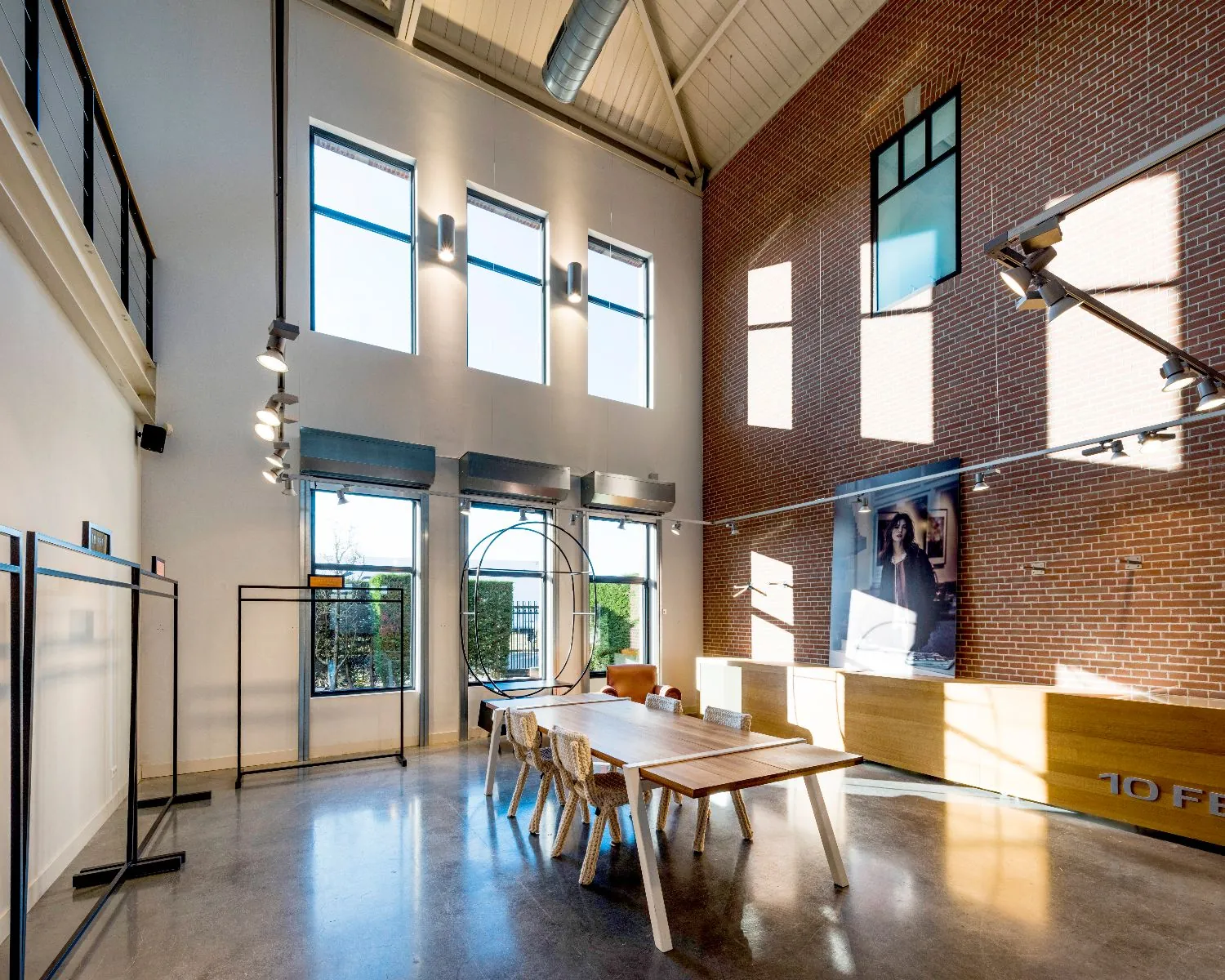 Een moderne, hoge ruimte met bakstenen muur, grote ramen, een houten tafel en een industriële inrichting.