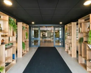 Ingang van een modern kantoorpand met houten open boekenkasten en groene planten aan weerszijden van de hal.