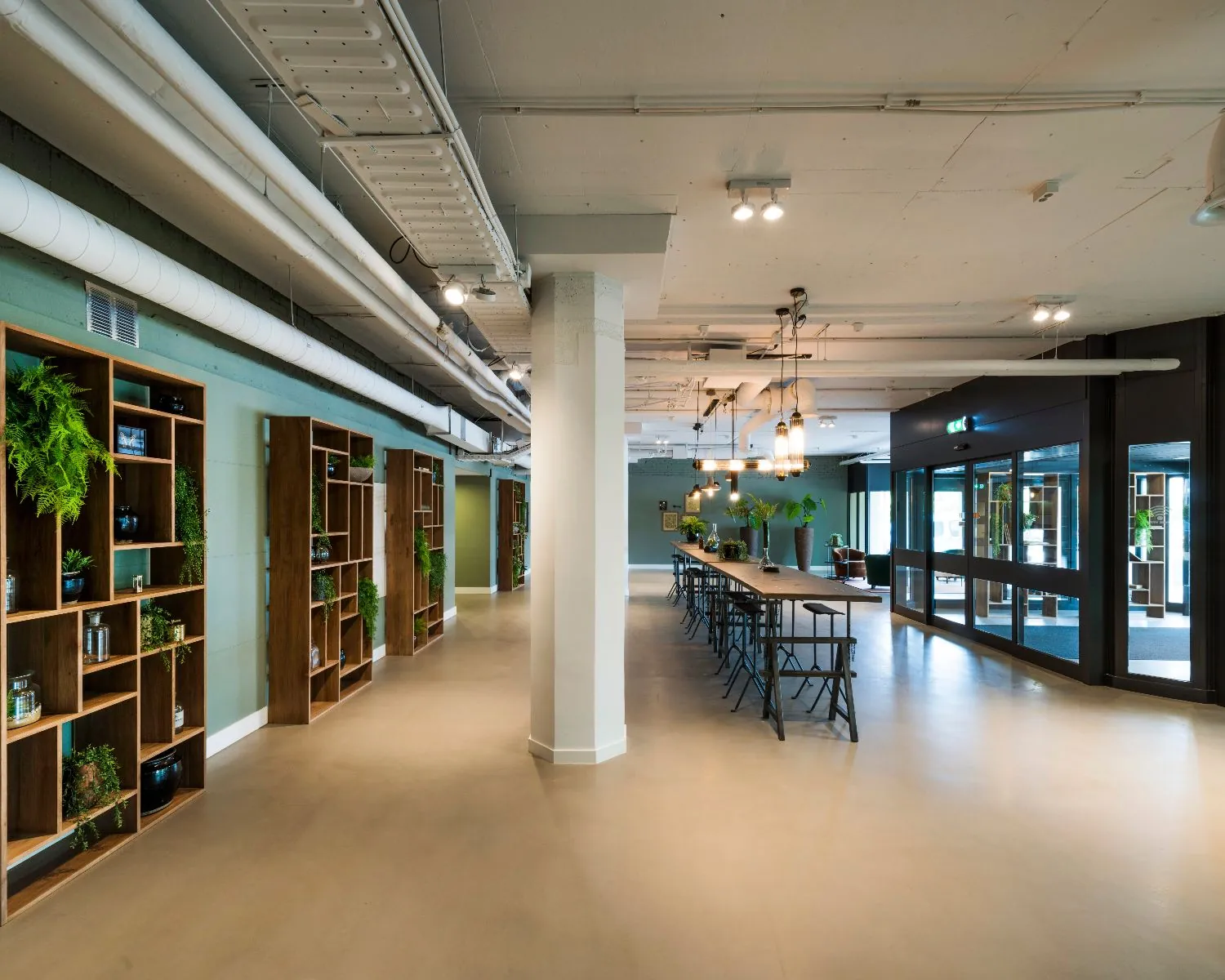 Modern kantoorinterieur met een lange vergadertafel, open boekenkasten met planten en industriële plafondleidingen.