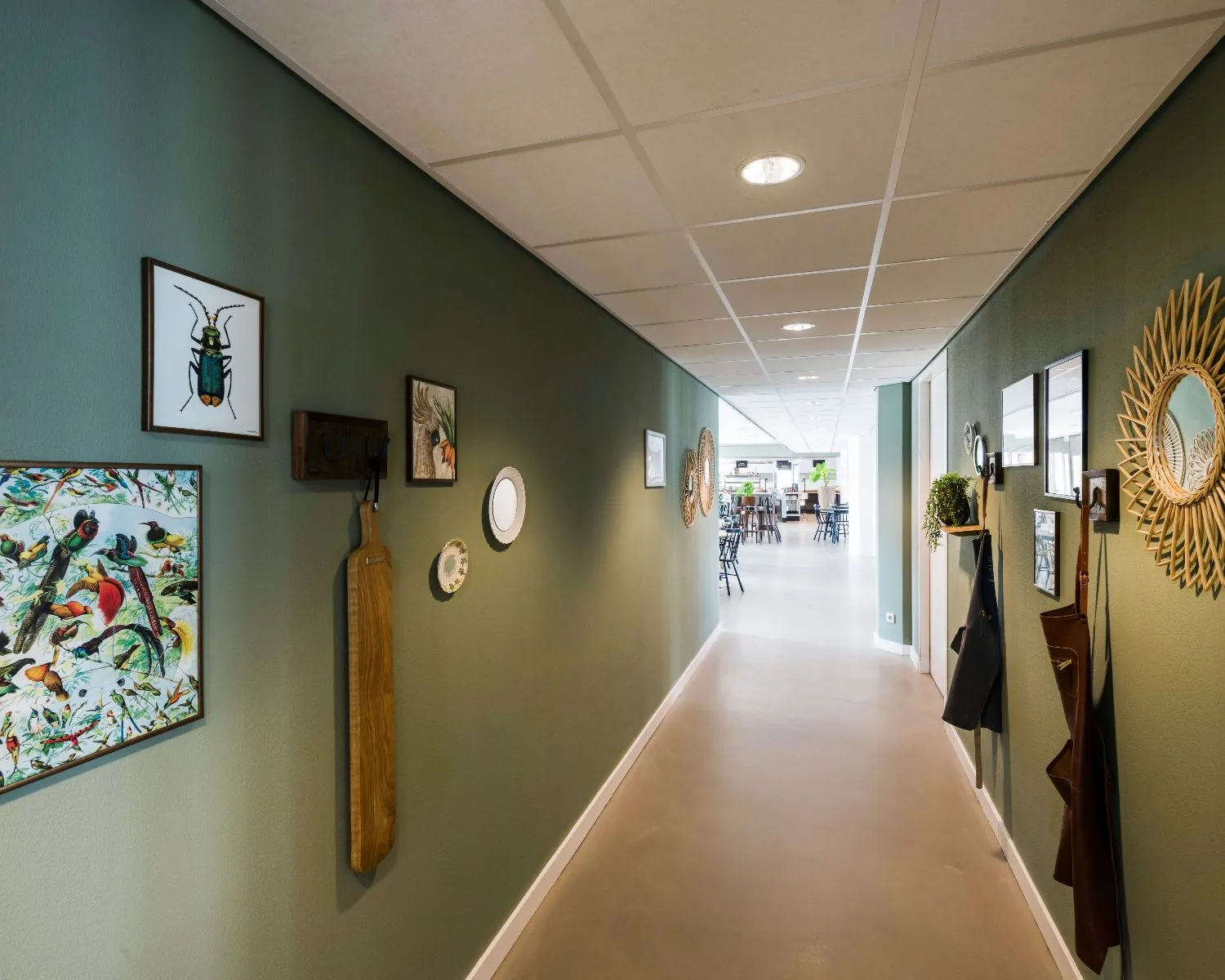 Lichte gang met groene muren, decoratieve wandkunst en een doorkijk naar een eetruimte met tafels en stoelen.