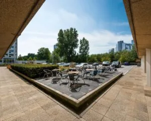 Terras met tafels en stoelen naast een parkeerplaats bij een kantoorgebouw in een stedelijke omgeving.