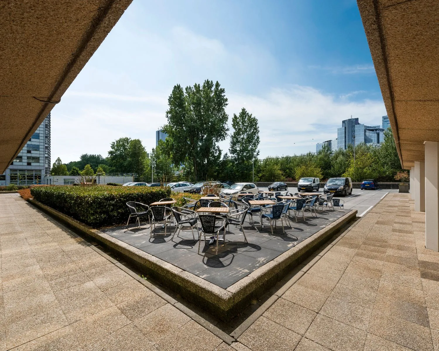 Terras met tafels en stoelen naast een parkeerplaats bij een kantoorgebouw in een stedelijke omgeving.
