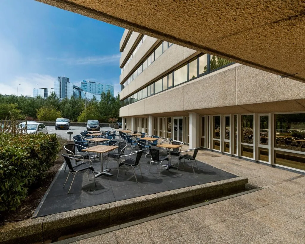 Terras met tafels en stoelen naast een modern kantoorgebouw met uitzicht op hoge gebouwen in de verte.