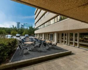 Terras met tafels en stoelen naast een modern kantoorgebouw met uitzicht op hoge gebouwen in de verte.